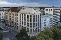 Flygfoto i fågelperspektiv över ett stadskvarter med en ljus hörnfastighet med valvbågar i gatuplan under en molnig skymningshimmel