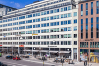 Byggnaden är en stor kontors- och affärsfastighet med en ljus fasad, troligen belägen i centrala Stockholm. I gatuplan syns flera kommersiella lokaler, varav några skyltar tydligt visar "DHL" och "Scandic Grand Central" samt "Brightby" på en intilliggande