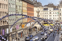 En livlig stadsgata med färgglada husfasader i grönt, gult, orange och beige. Tvärs över gatan hänger bågar dekorerade med blå och lila blommor. Bilar, cyklar och människor rör sig längs gatan, medan skyltar och butiker kantar trottoarerna.