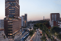 En arkitektonisk visualisering av en modern skyskrapa i tegel som reser sig över en livlig stadsmiljö i skymning. Vid byggnadens fot syns spårvagnar, bussar och biltrafik, omgivna av lummiga trädalléer och intilliggande bostadshus.