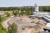 En översiktsbild av en vit låg byggnad, med ett tillhörande högre torn. I mitten av bilden syns en grusplan och en mindre gul traktor som schaktar grus.