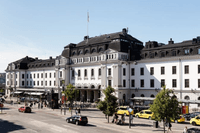 Exteriör av Stockholms centralstation en solig dag med bilar, taxibilar och fotgängare på gatan framför den vita byggnaden.