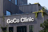 Exteriör av GoCo Clinic. Den moderna byggnaden har en fasad av lodräta träribbor och stora vita bokstäver som bildar namnet. På takterrasserna syns gröna växter och hängande klätterväxter mot en klarblå himmel.