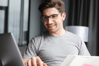 En leende ung man med glasögon som sitter vid en bärbar dator och studerar eller arbetar med en bok framför sig.