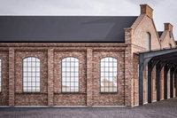 En lång industribyggnad i rött tegel med karaktäristiska bågfönster. Byggnaden har ett mörkt tak och en modern utbyggnad i form av ett svart stålskelett som skapar en övertäckt gångväg längs med fasaden. Markytan består av mörk gatsten.