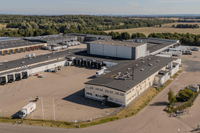 Flygfoto i fågelperspektiv över ett stort logistik- och lagerkomplex med lastkajer, lastbilar och omgärdande åkermark.