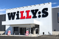 Exteriör av en Willys-butik med den klassiska logotypen på en vit fasad ovanför entrén. Till höger syns skyltar för Svenska Spel, DHL och uttagsautomat, och framför ingången finns parkerade cyklar under en klarblå himmel.