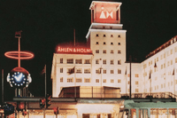 Klassiskt nattfoto på Åhlén & Holms byggnad med upplysta skyltar och ett torn. I förgrunden syns en ljusinstallation och en spårvagn.