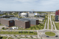 En vy från ovan över ett modernt kontorsområde med varierad arkitektur och mycket grönska. I förgrunden löper en väg där en grön spårvagn passerar en rondell. Till vänster syns stora bruna tegelbyggnader med solpaneler på taken, och i bakgrunden sticker e