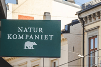 Grön butiksskylt med texten NATUR KOMPANIET och en björnlogotyp som hänger ut från en fasad