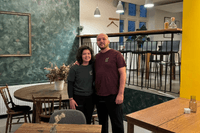 En kvinna med mörkt lockigt hår och en man i en vinröd t-shirt står tillsammans i en restaurang- eller caféinteriör. De står framför en mörkgrön, mönstrad fondvägg med en tom tavelram och ett runt träbord med stolar. Till höger bryter en starkt gul vägg a