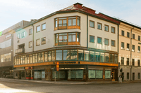 Hörnfastighet i tre våningar med ljus puts, trädetaljer och skyltfönster vid en korsning under en solig dag.