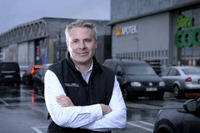 En man i vit skjorta och svart väst står med armarna i kors på en parkeringsplats framför en handelsplats. I den suddiga bakgrunden syns parkerade bilar och butikskedjor som "Kronans Apotek" och "Stora Coop". Det är en mulen dag och marken är blöt av regn