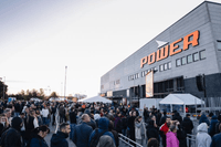 En stor folkmassa köar utanför en stor Power-elektronikbutik under dagsljus. Människor står tätt i fållor avgränsade med metallstängsel framför den grå butiksbyggnaden med den orangea Power-logotypen.