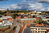 Flygfoto i fågelperspektiv över ett stadslandskap med kyrka, tegelbyggnader med solceller och skogsklädda höjder i bakgrunden under en dramatiskt molnig sky.