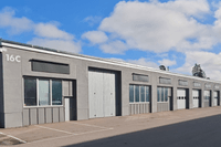 En lång, grå industrifastighet i en våning med flera sektioner under en blå himmel med lätta moln. Byggnaden har stora fönsterpartier, industriportar och en markerad husnummering "16C" på hörnet. Framför byggnaden finns en asfalterad yta med parkeringsmar