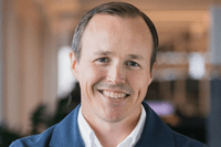 Ett närporträtt av en leende man med kort ljusbrunt hår. Han bär en vit skjorta under en blå, texturerad kavaj eller kofta. Bakgrunden är ljus och mjukt suddig, vilket ger bilden en professionell känsla.