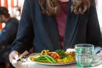 Närbild av en person som bär en bricka med en måltid bestående av ris, grönsaker samt ett glas vatten i en restaurangmiljö.