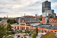 En vid vy över Sickla i Nacka där historiska tegelbyggnader möter modern arkitektur. I förgrunden syns Dieselverkstaden och Filmstaden vid ett torg med uteserveringar och grönska, medan den höga profilen av Atlas Copco-skrapan dominerar bakgrunden mot en 