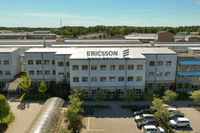 Flygfoto över Ericssons stora anläggning i ljus fasad med parkerade bilar och grönområden under en klarblå sky.