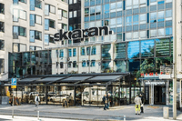 Exteriörbild i ögonhöjd över entrén till gallerian och skyskrapan Skrapan med inglasad uteservering, fasadskyltar och fotgängare.