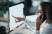 En kvinna med glasögon sitter vid ett skrivbord, talar i en smartphone och pekar på en datorskärm som visar ett kalkylark eller tabell.