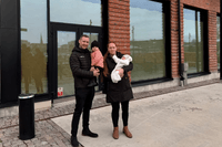 En man och en kvinna står utomhus framför en modern tegelbyggnad med stora glaspartier. Båda håller i varsitt barn, varav ett är en baby i en vit overall. De är klädda i mörka vinterjackor och miljön ser ut som ett nybyggt område med stenlagd mark.