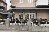 Fasaden till ett bageri, konditori och café med en uteservering i förgrunden, prydd med blomlådor fyllda med färgglada penséer.