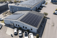 Flygfoto över en industrifastighet med stora solcellspaneler installerade på taken. Byggnaden omges av en parkeringsplats med bilar och ligger i anslutning till ett grönområde.