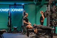 En kvinna gör hantelpress på en träningsbänk i ett gym med turkosa väggar och neonskylten "POWER CORNER".