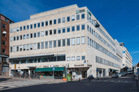 En femvånings ljus stenbyggnad i ett gatuhörn under klarblå himmel. På bottenvåningen finns flera butiker, bland annat en livsmedelsaffär med grönt markistak och en butik med skylten "Kreatiffa". Ovanför syns många rektangulära fönster i rad längs fasaden