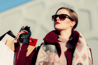 En elegant kvinna i solglasögon, rött läppstift och mörkröd jacka som bär på flera shoppingkassar utomhus.