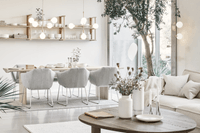 Ett ljust och modernt vardagsrum med skandinavisk design. I förgrunden står ett runt träbord med vita vaser och torkade blommor. Till höger syns en beige soffa med ljusa kuddar och bakom den ett stort grönt träd intill fönstret. I bakgrunden finns ett mat
