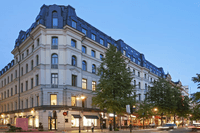 En ståtlig hörnfastighet i klassisk stil under blå timmen, belägen vid en livlig stadsgata med träd och gatulyktor. Byggnaden har en ljus putsad fasad, ett mörkt mansardtak och butiker med upplysta skyltfönster i gatuplan. Bilar med ljusspår rör sig på vä