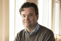 Ett porträtt av en man med mörkt, vågigt hår, klädd i en ljusblå skjorta under en brun stickad tröja. Han tittar direkt in i kameran med ett neutralt uttryck. Bakgrunden är ljus och mjukt suddig med vertikala linjer från fönsterpartier som släpper in natu