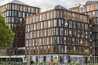 Stora, moderna kontorsbyggnader med rundade hörn och en karaktäristisk fasad av brun metall och vertikala glaspartier. I förgrunden syns en stadsmiljö med en gata, en spårvagn, en cyklist och gröna träd under en växlande molnighet.