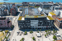 Flygfoto över ett torg och stadsområde med en stor mörk fastighet utrustad med solpaneler och växttak vid havet.