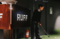 Närbild på en skylt med texten "RUFF" i en inomhusmiljö för golf, med en person som skymtas i bakgrunden när de svingar en golfklubba på en konstgräsmatta.