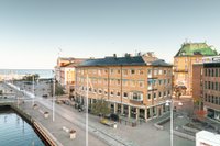 En vy över en stadsbild vid vattnet i mjukt dagsljus. I centrum står en hörnbyggnad i gult tegel med fyra våningar och skyltar för restaurangen 'Frimans' samt kontoret 'Ludvig & Co'. Till vänster löper en stenlagd kaj längs ett vattenparti, och i bakgrund