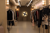 En exklusiv klädbutik med industriell känsla och varm belysning. Klädställningar med herrkläder kantar väggarna i en avlång lokal med synliga rör i taket, och längst in i rummet sitter en stor, belyst fläkt som en dekorativ detalj.