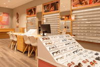 Interiörbild i ögonhöjd från en optikerbutik med glasögonställ, provbord, stolar och en kassadisk i trä.