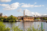 En vy över ett vattennära industriområde med äldre tegelbyggnader och en hög skorsten under en blå himmel med vita moln. I förgrunden syns glittrande vatten och gröna träd som ramar in bilden.