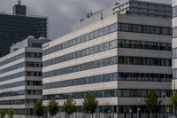 En vy över moderna kontorsbyggnader i ett industriområde under en mulen himmel. I förgrunden dominerar en vit flervåningsbyggnad med ett mönster av små cirklar på fasaden och 'Ericsson'-logotypen synlig uppe till höger. I bakgrunden syns en mörkare, hög s