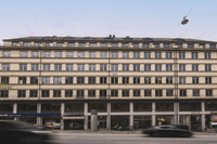 En bred fasadbild av en stor kontorsbyggnad i flera våningar vid en trafikerad stadsgata. I gatuplan syns butiker och verksamheter som Visita, Fabrique och ett apotek. Suddiga bilar i rörelse passerar framför byggnaden under en klar himmel.