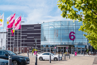 Väla köpcentrums entré nummer 6 med en stor rundad glasfasad, flaggor som vajar i vinden och parkerade bilar i förgrunden.