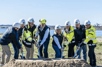 En grupp människor i byggvästar och skyddshjälmar tar ett gemensamt första spadtag för ett byggprojekt. De står utomhus vid en sandhög med havet i bakgrunden under en klarblå himmel.