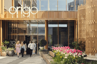 En modern byggnad med namnet "Origo" ovanför entrén. Fasaden är klädd i ljusa träribbor och stora glaspartier. I förgrunden syns en stenlagd gångväg kantad av blommande rosa och vita tulpaner, och en grupp människor är på väg in i byggnaden.