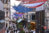 En vy över en tät stadsgata/gågata i skymningen eller på kvällen, dominerad av byggnader med äldre arkitektur och moderna inslag. Gatan är dekorerad med stora, spända tygsegel i blåa, röda och lila nyanser som hänger över gångvägen. Längre fram i bilden s