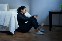 En mörk inomhusbild visar en kvinna i svarta, åtsittande kläder sittande på ett trägolv med korslagda ben, lutad mot en säng med vitt täcke. Hon håller ett glas vatten och ser tankfull ut. I det svagt upplysta rummet syns ett nattduksbord med böcker och e