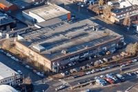 Flygfoto över en lång tegelbyggnad i ett industriområde. Framför byggnaden finns en stor parkering fylld med bilar, och gatorna runtomkring kantar av parkerade fordon.